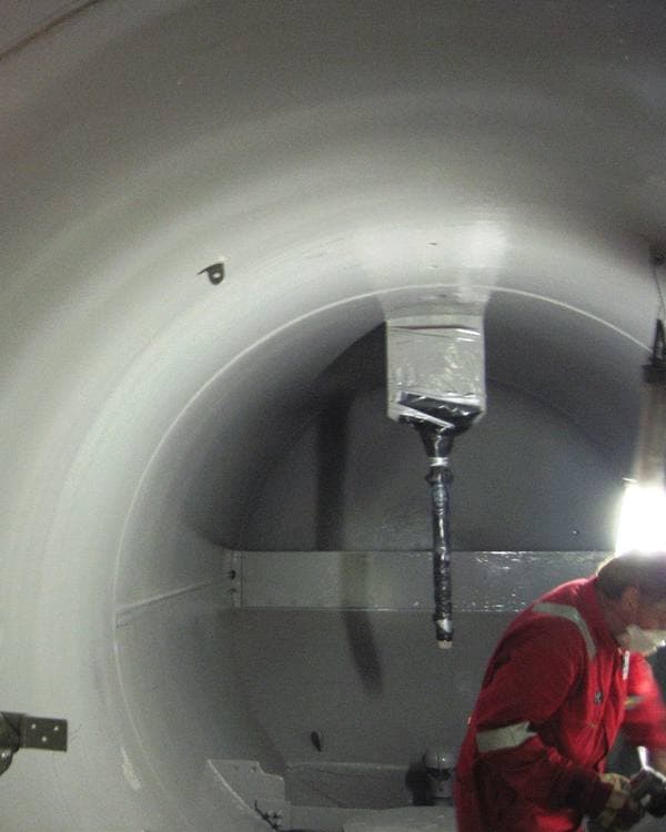 Worker in red coveralls and a dust mask inside a large cylindrical vessel that has been freshly coated pale gray